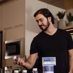 Man in a modern kitchen holding a vitamin supplement bottle, surrounded by health products.